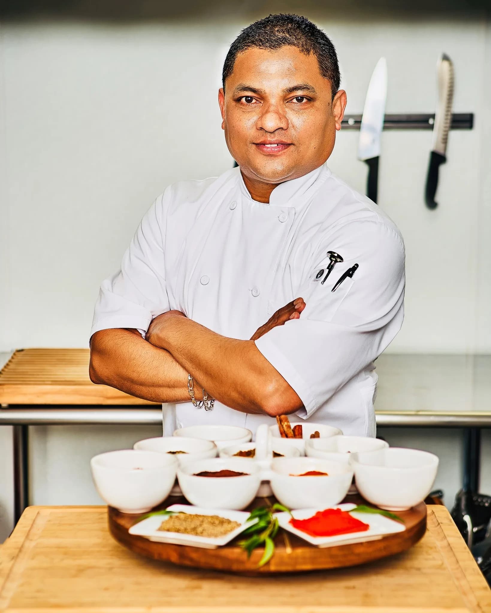 Chef Kal Peiris standing confidently with arms crossed behind bowls of traditional Sri Lankan spices